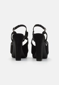 Sorte semskede høyhælte sandaler med åpen bak, justerbare ankelstropper og en grov hæl. Teksturert materiale med glatte accenter.