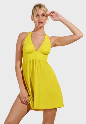 Donna che indossa un vestito giallo senza maniche con scollatura a V profonda, posando con un braccio alzato e l'altro che tiene l'orlo del vestito.