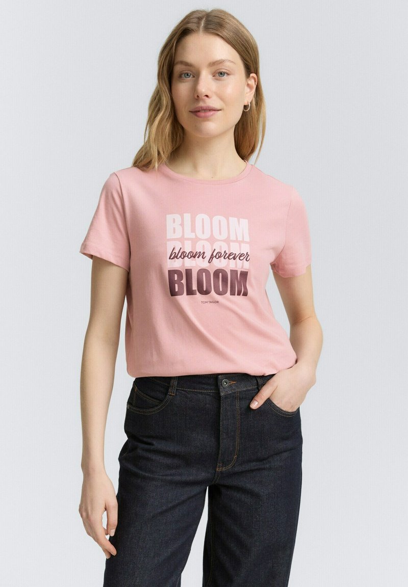 Camiseta de algodón rosa claro con mangas cortas, que presenta el texto "BLOOM" en diferentes colores y tipografías. Combinada con jeans oscuros.