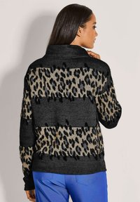 Frau mit langen dunklen Haaren, die einen langärmeligen schwarzen und beigen Leopardenmuster-Pullover und leuchtend blaue Hosen trägt, seitlich vor einem neutralen Hintergrund steht.