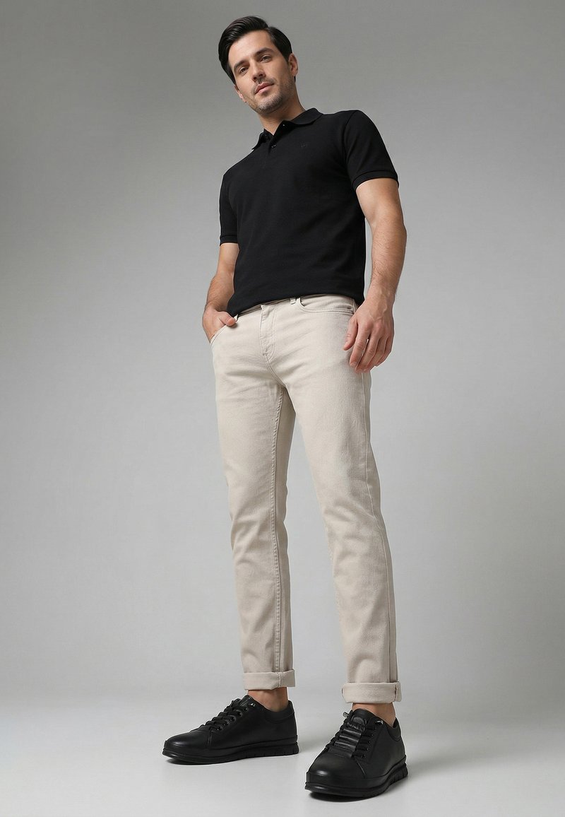 Mann trägt schwarzes Poloshirt, beige hochgekrempelte Hose und schwarze Sneaker, steht mit Händen in den Taschen vor grauem Hintergrund.