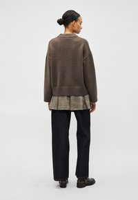 Γυναίκα με τα μαλλιά σε κότσο, φοράει φαρδιά καφέ μπλούζα πάνω από πουκάμισο με καρό πλισέ, σκούρο φαρδύ παντελόνι και μαύρα μποτάκια μέχρι τον αστράγαλο, στέκεται γυρισμένη προς τα πίσω.