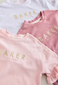 Kolm puuvillast T-särkki: valge, helesinine ja tumepink. Igaühel on kuldne "BAKER" logo. Heleda roosa särgil on lainelised varrukad.