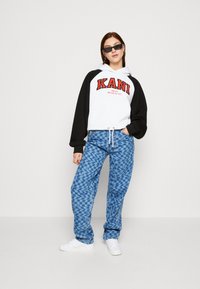 Hanorac cu glugă, cu mâneci negre și corp alb, având logo-ul roșu "KANI". Blugi largi cu un model albastru și adidași albi.