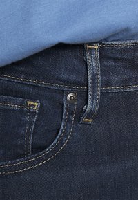 Acercamiento de un bolsillo de jeans de mezclilla azul con costuras amarillas y una camisa azul clara metida encima de la cintura.