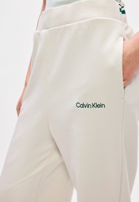 Ανοιχτό καφέ φόρμες με ελαστική μέση, πλαϊνές τσέπες και κεντητό λογότυπο "Calvin Klein" σε σκούρο πράσινο στο αριστερό μηρό. Λείο υφάνισμα.