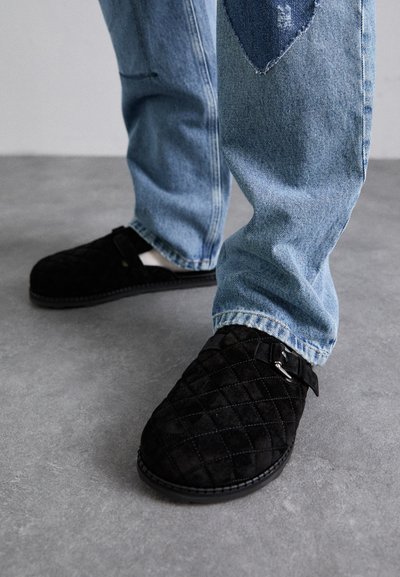 Black suede slip-on shoes with a quilted pattern and buckle detail, paired with light blue distressed denim jeans.