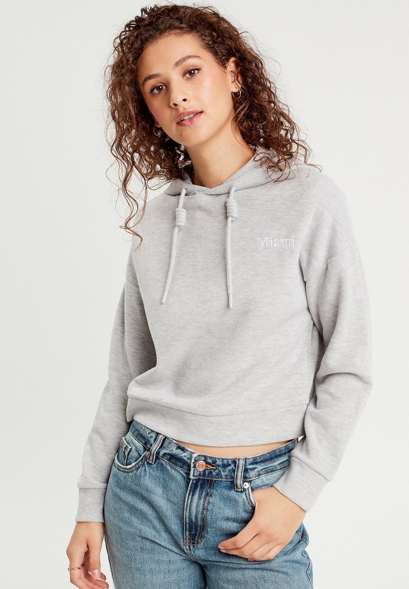 Lichtgrijze cropped hoodie met trekkoorden, voorzien van een eenvoudig ontwerp en "Miami" geborduurd op de borst. Gdraagd met hooggesneden denim jeans.