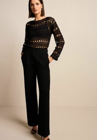 Schwarzes, gemustertes Strickoberteil mit langen Ärmeln, kombiniert mit hochgeschnittenen schwarzen Hosen. Das Model steht mit den Händen in den Taschen; minimalistische Accessoire-Details.