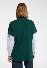 Zielony top z imitacją kołnierzyka, krótkimi rękawami i luźnym krojem. Nałożony na jasnoniebieską koszulę z długim rękawem, o fakturowanej powierzchni.