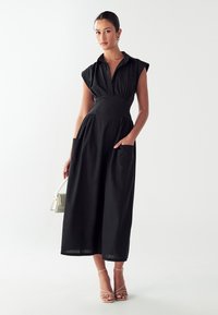 Črna srajčna obleka s kratkimi rokavi, s prilegajočim pasom in dvema sprednjima žeparjema; izdelana iz lahkega materiala, sega do sredine meč.