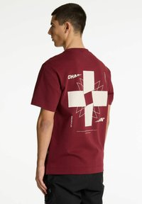 Bordo medvilninis marškinėliai su dideliu baltu grafinio dizaino spaudiniu ant nugaros, įskaitant kryžiaus formą ir geometrinius raštus, trumpomis rankovėmis.