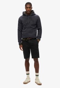Donkergrijze hoodie met logo, gelaagd over een geruit shirt. Zwarte shorts, crèmekleurige sneakers en witte sokken met groene en rode strepen.