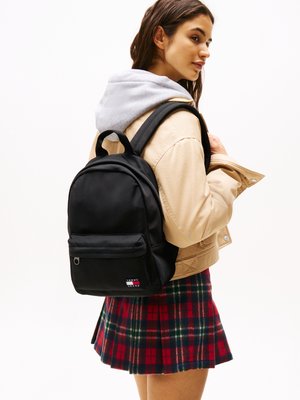 Young woman wearing a beige jacket, gray hoodie, red plaid skirt, and carrying a black Tommy Jeans backpack over one shoulder.