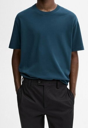 T-shirt en coton couleur bleu sarcelle à col rond avec manches courtes, associé à un pantalon foncé. Le t-shirt présente une coupe décontractée et un design simple, sans motifs visibles.