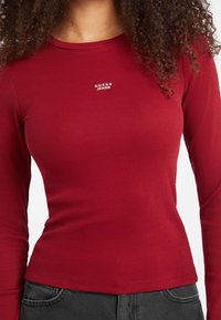 Top lungo a maniche rosse con scollatura a girocollo e un piccolo logo bianco "GUESS JEANS" sul petto. Silhouette aderente, texture morbida.
