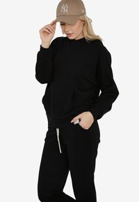 Czarna bluza z kapturem z kieszenią z przodu, w połączeniu z czarnymi spodniami dresowymi. Całość dopełnia beżowa czapka z daszkiem z białymi akcentami logo.