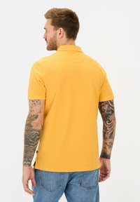 Geel poloshirt met korte mouwen, een gladde katoenen textuur en zijsplitten. Kenmerken zichtbare tatoeages op de armen en een standaard kraagontwerp.