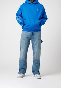 Modrá mikina s kapucí s logem na levé straně hrudi a bílým nápisem na rukávu, doplněná o světlé modré džíny s nákladními kapsami a bílé tenisky.