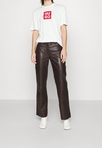 Camiseta blanca de manga corta con el logo rojo de Hugo, combinada con pantalones de cuero marrón oscuro y botas blancas hasta el tobillo. Diseño sencillo y textura suave.