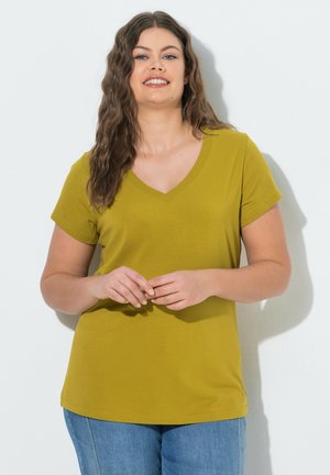 Frau mit langen, welligen Haaren trägt ein senfgelbes kurzärmeliges V-Ausschnitt-Shirt und blaue Jeans und steht vor weißem Hintergrund.