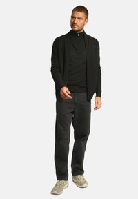 Czarny kardigan na zamek błyskawiczny noszony na czarnym swetrze, połączony z ciemnymi dżinsami i jasnymi trampkami. Kardigan ma luźny krój i ściągacze.