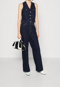 Jeansowa kamizelka w ciemnym niebieskim kolorze z frontowymi guzikami i dwoma kieszeniami na piersi, zestawiona z szerokimi spodniami jeansowymi w ciemnym niebieskim odcieniu oraz czarno-białą torebką.