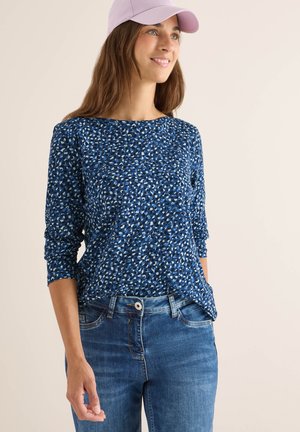 Blå mönstrad långärmad topp med en avslappnad passform, ihop med mellanhöga blå jeans. En ljuslila keps fulländar outfiten.
