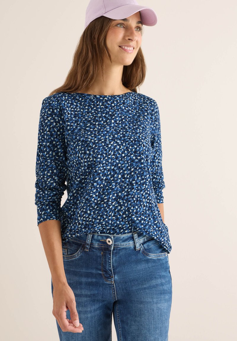 Blau gemusterte Langarmoberteil mit lockerer Passform, kombiniert mit mittel-hoch geschnittenen blauen Jeans. Eine helllila Mütze rundet das Outfit ab.