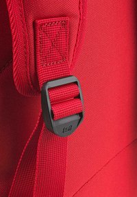 Mochila de tela roja con un broche de plástico negro, correa de cinta gruesa y superficie texturizada con detalles de costura reforzada.