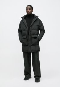 Črni podloženi plašč z visokim ovratnikom, sprednjo zadrgo in več žepi. Nošen preko črnega puloverja z visokim ovratnikom in kombiniran z ohlapnimi črnimi hlačami.