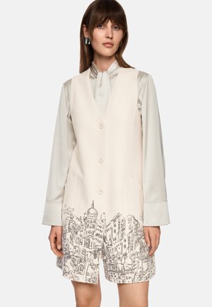 Beige Weste mit einem Motiv von Stadtlandschaften am Saum, featuring einen V-Ausschnitt, drei Knöpfen und seitlichen Taschen. getragen über einer hellen Bluse.
