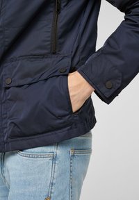 Marinblå isolerad jacka med dragkedja framtill, stor ficka med tryckknapp och texturerat tyg. Kombineras med ljusblå denim.