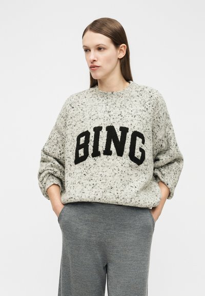 Frau mit glatten braunen Haaren trägt einen grau gesprenkelten Pullover mit dem schwarzen Schriftzug "BING" und lockere graue Hosen, Hände in den Taschen.