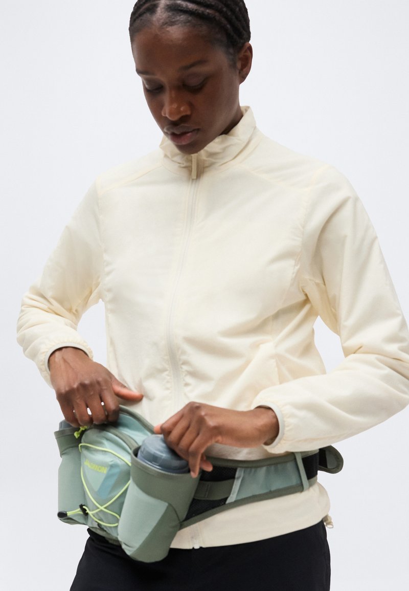 Femme ajustant une banane bleu clair avec des bouteilles d'eau, portant une veste à fermeture éclair crème et un pantalon noir, sur un fond uni.