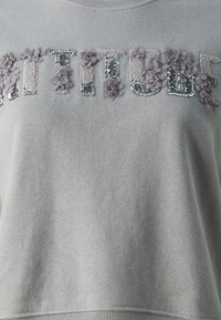 Sudadera gris claro con tejido texturizado, aplicados florales y letras con lentejuelas que dicen "ACTITUD". Cuello redondo y corte relajado.