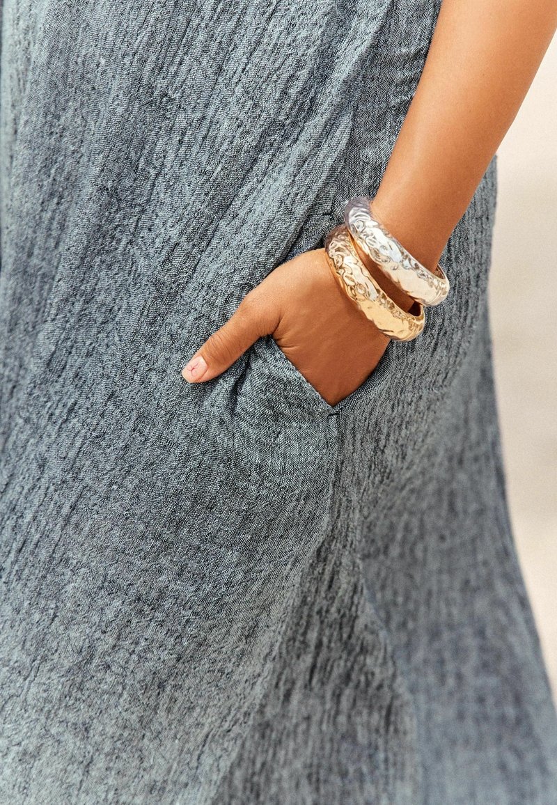 Main portant deux bracelets métalliques texturés reposant dans la poche d'une robe grise en tissu froissé.