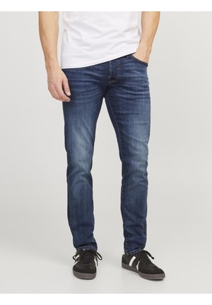 Jeans de mezclilla azul oscuro con un ajuste slim. Presentan un desvanecimiento sutil, diseño de cinco bolsillos y costuras en contraste. Combinados con zapatillas negras.