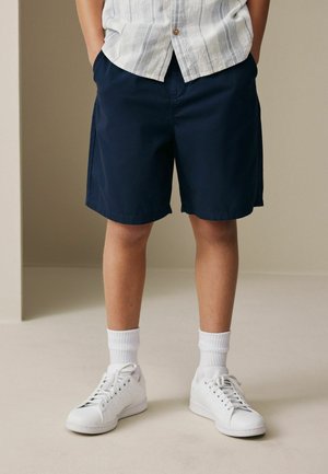 Child wearing navy blue shorts, white sneakers, white socks, and a light-colored striped shirt with hands in pockets, standing indoors.