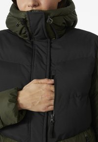 Chaqueta de pluma negra y verde oliva con un alto cuello, bolsillo con cremallera lateral y texturas contrastantes. Presenta un ajuste ceñido y capucha.