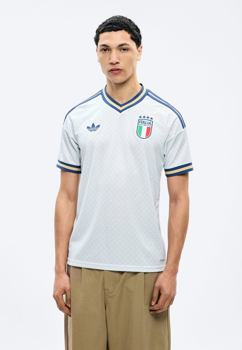 Mladý muž v bielom futbalovom drese Talianska s modrým a zlatým lemovaním a béžovými nohavicami, stojaci na jednoduchom svetlom pozadí.