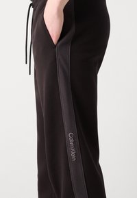 Melni sporta bikses ar sānu paneli tumšākā tonī, ar kabatu un izšūtu "Calvin Klein" logo pelēkā krāsā. Mīksts audums, brīvs piegriezums.