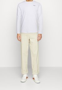 T-shirt bianco a maniche lunghe con un piccolo logo, abbinato a pantaloni color crema e sneakers bianche con accenti arancioni.