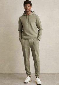 Olijfgroene sweatshirt met capuchon en trekkoorden, gecombineerd met bijpassende taps toelopende broeken. Afgemaakt met witte sportsneakers. Soepele stof.