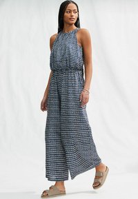 Marinblå och vit mönstrad jumpsuit med hög halsringning, samlad midja och löst sittande vida ben. Buren med neutrala sandaler.