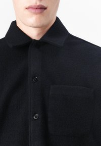 Black textured shirt with a button-up front, collared neck, and a chest pocket. Made of a soft fabric with a matte finish.