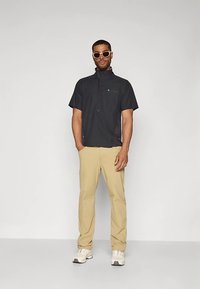 Mężczyzna w beżowych spodniach, czarnej bluzie z krótkim rękawem i zamkiem, białych tenisówkach oraz jasnych okularach przeciwsłonecznych, stojący na tle jednolitej białej ściany.