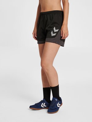 Persoon draagt zwarte sportshorts met grijze bies en blauwe sneakers met witte chevron-logo's, staat met één voet iets naar voren.