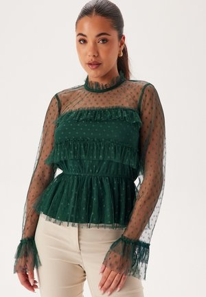 Blusa verde con volant sovrapposti, maniche lunghe trasparenti e motivo a pois. Presenta un collo alto e un tessuto strutturato.