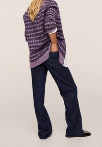 Osoba v tmavě modrých širokých džínách a svetr s fialovým vzorem, se rukou v zadní kapse, postávající na neutrálním pozadí.